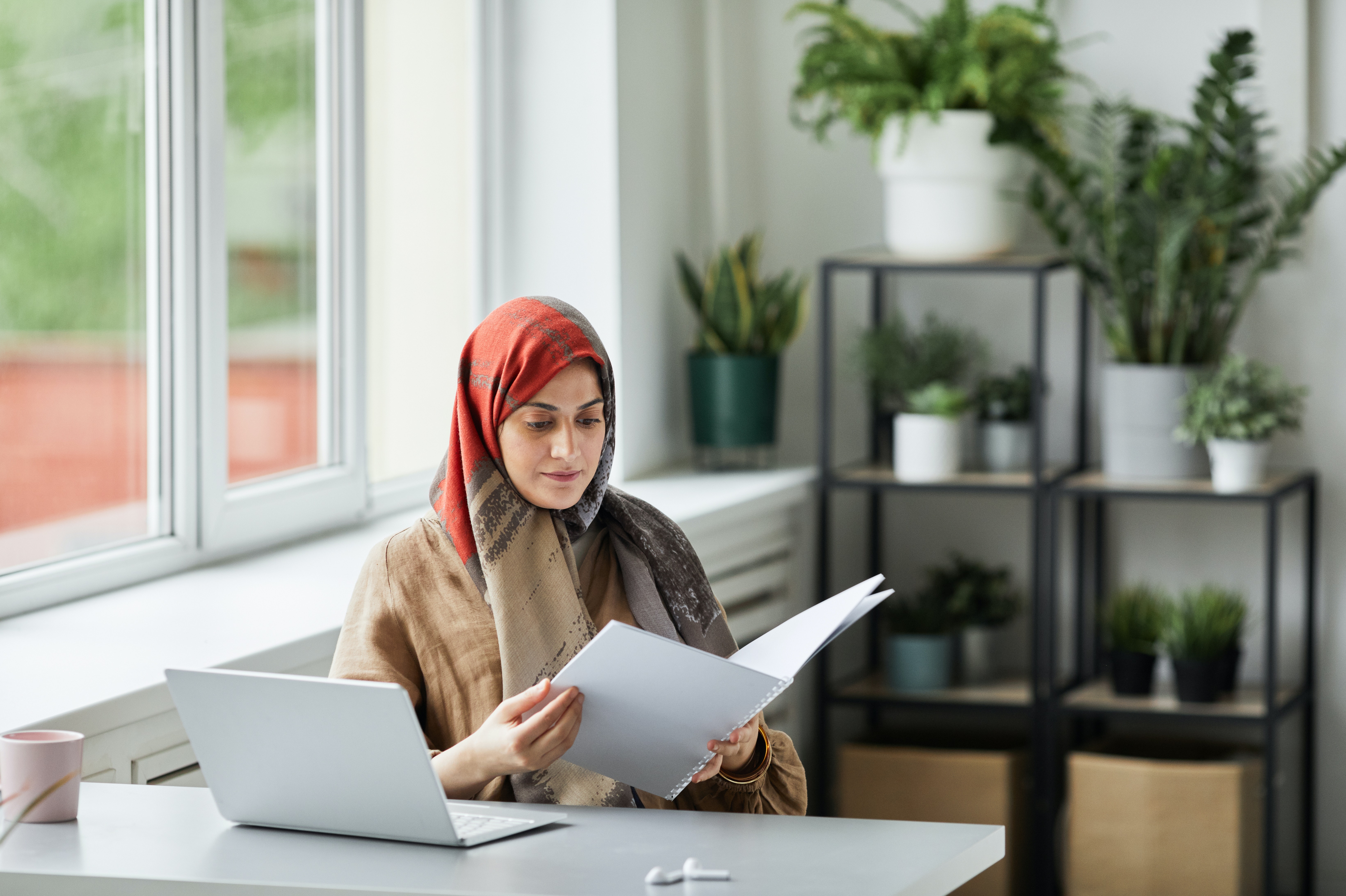 Lady with headscarf looking at booklet with laptop in front of her