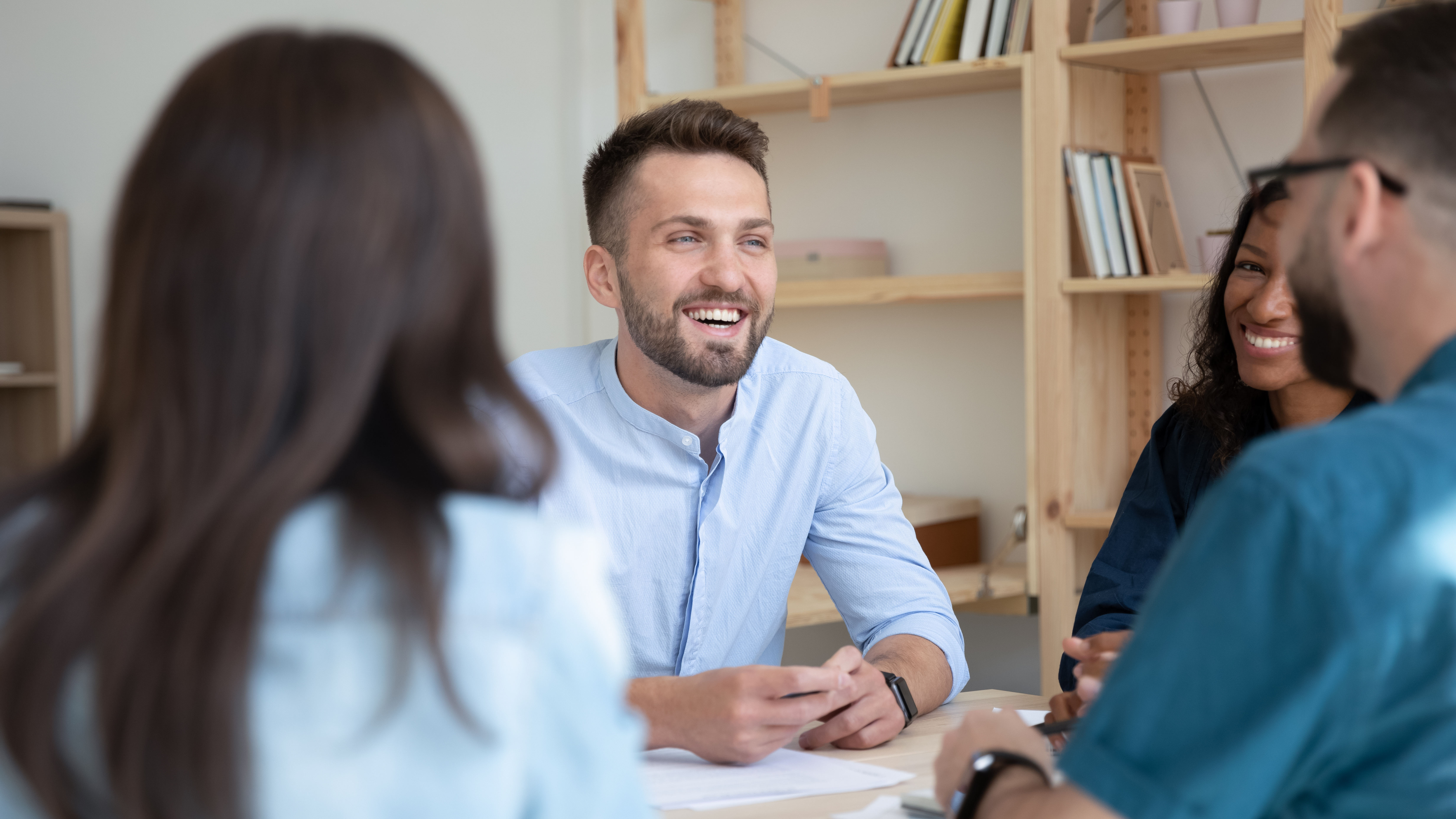 Male colleague smiling and in focus with other colleagues sat around table