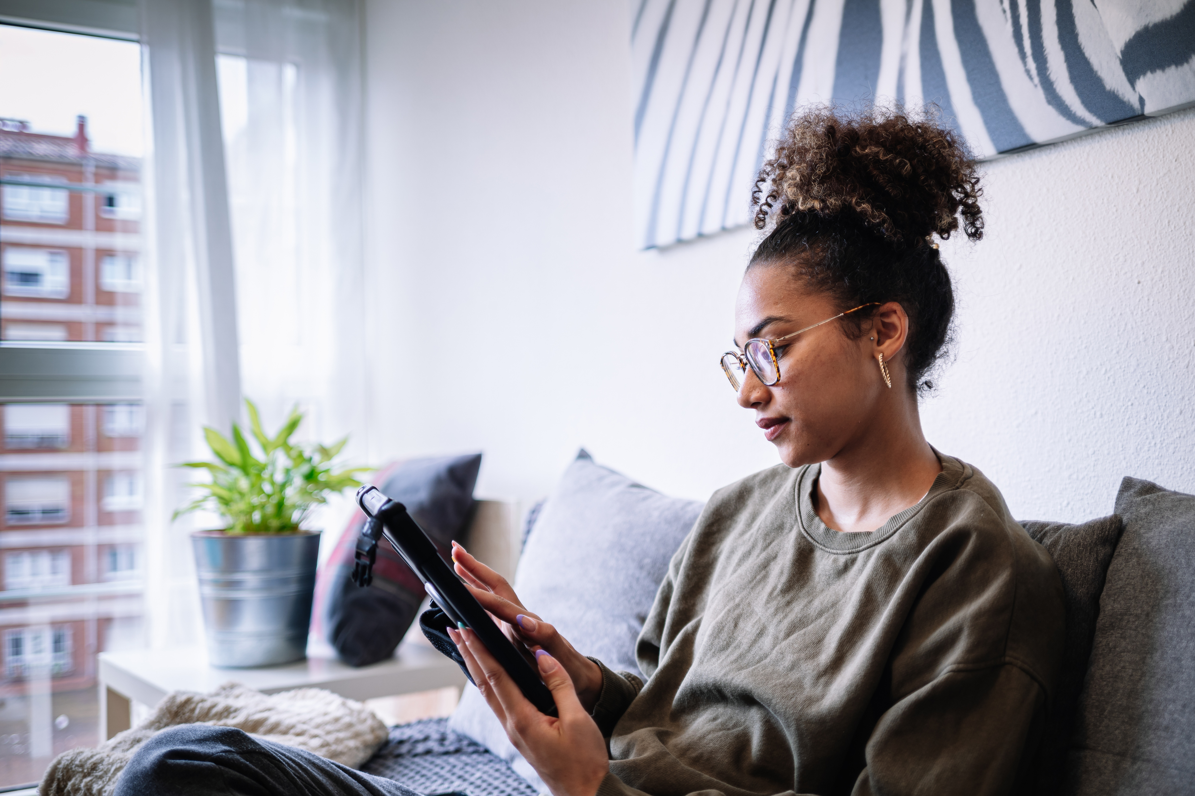 Young woman sat on sofa near window in high-rise apartment looking at tablet device