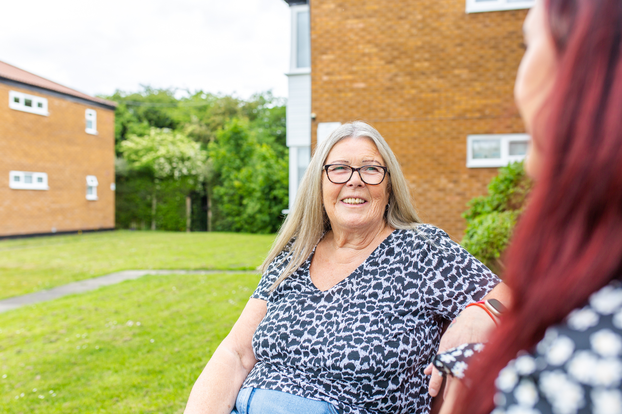 Older woman sat down, facing camera, talking to woman with red hair