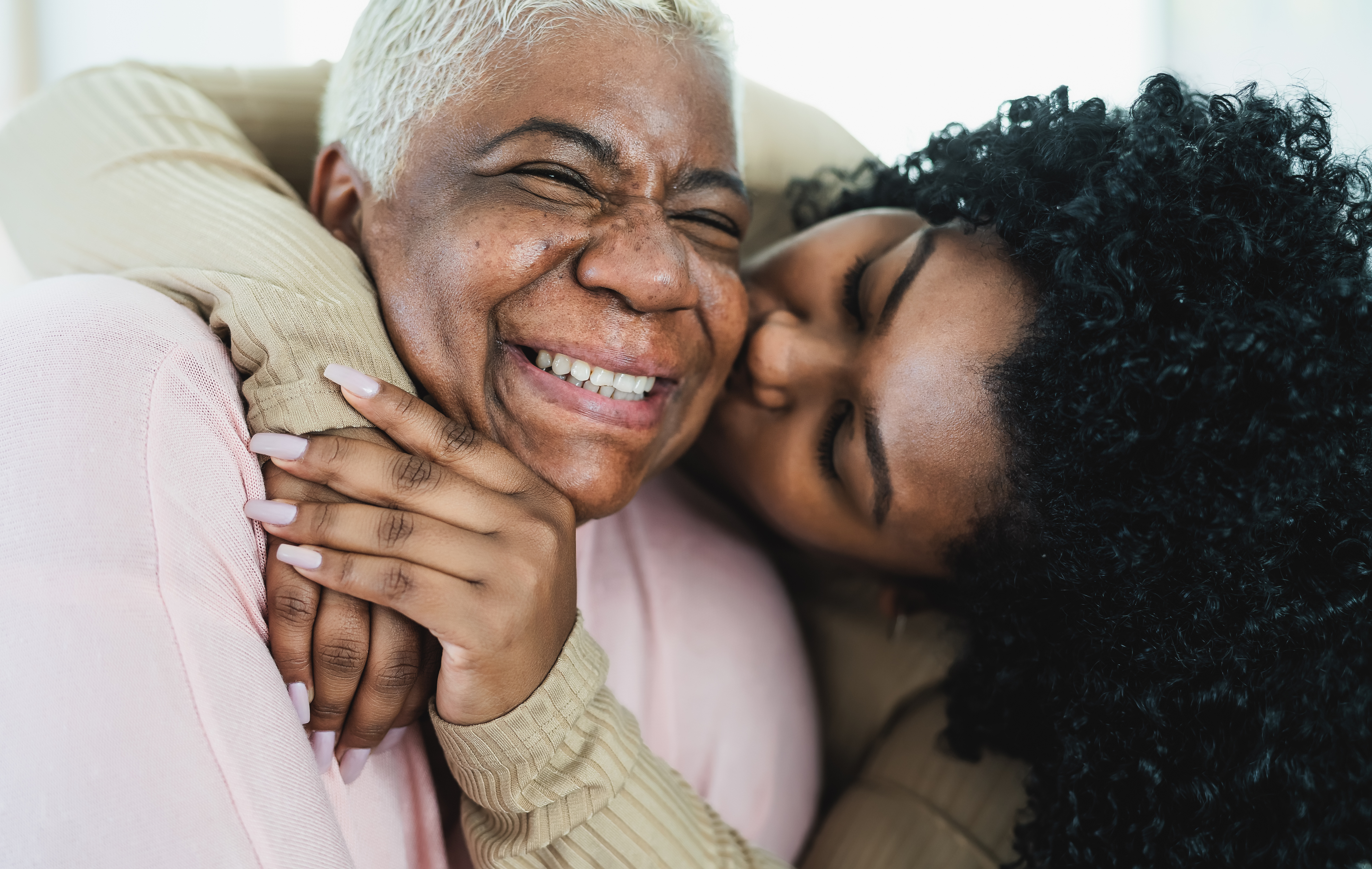 Close-up of smiling older woman in pink top being hugged tightly by smiling young woman in beige top