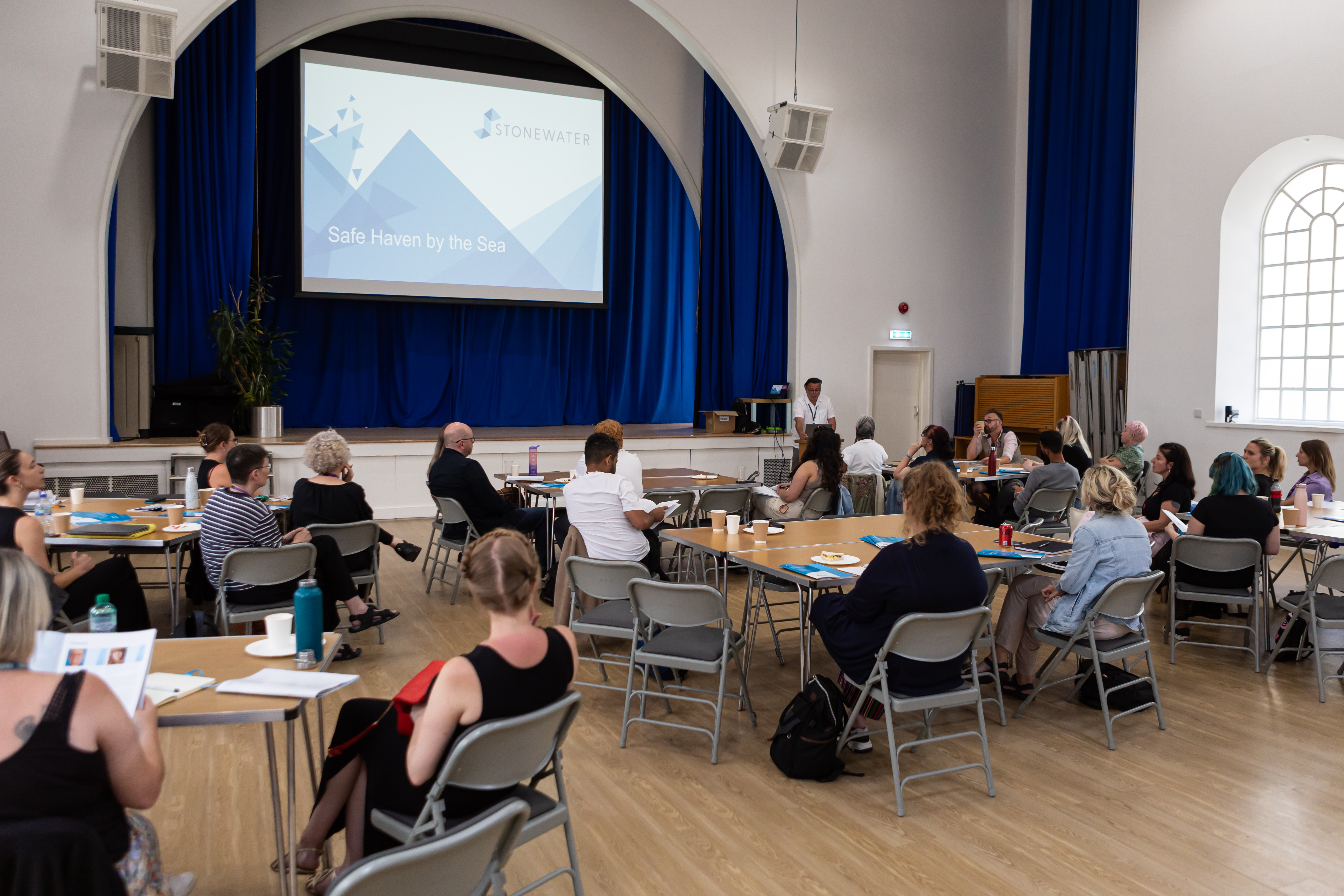 People are sat around tables in a large hall facing a stage where a presentation is being held.